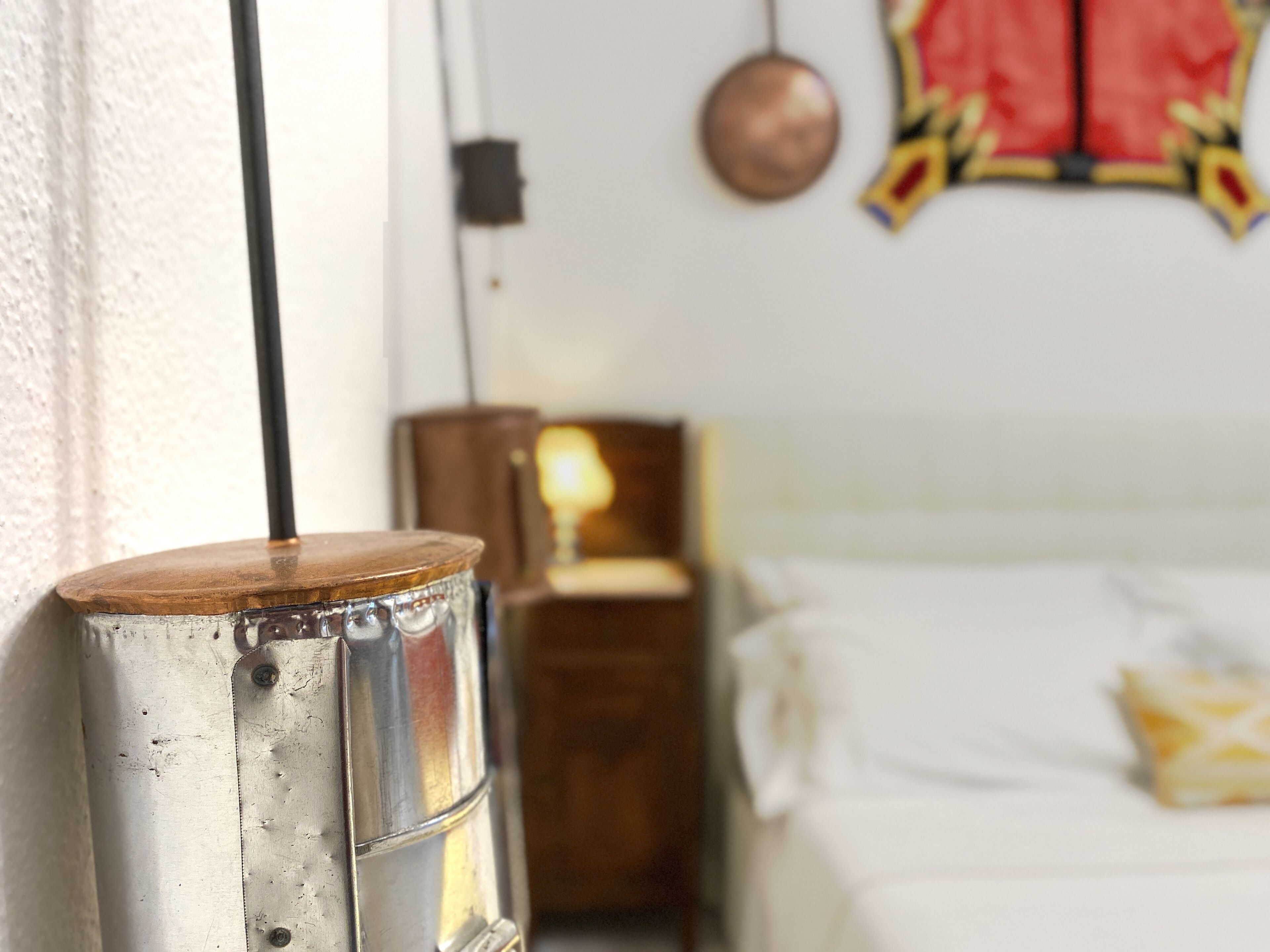 A metal radiator with a wooden top, a wooden nightstand with a lamp, and a bed with white bedding and a yellow pillow in a room with a white wall and a decorative hanging ornament.