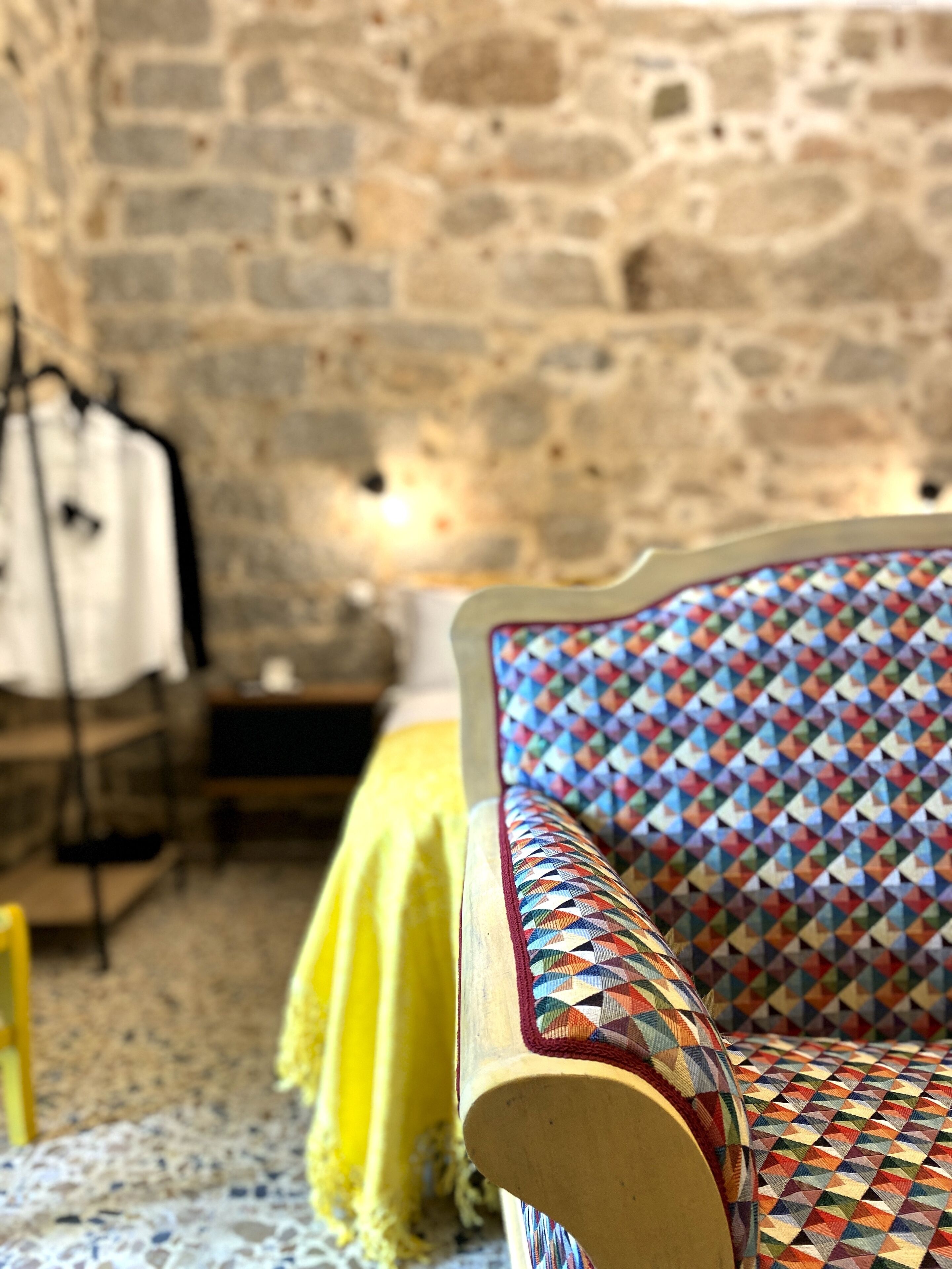 A room with a stone wall, a bed with a colorful geometric-patterned headboard, a yellow bedspread, and a white shirt hanging on a rack.