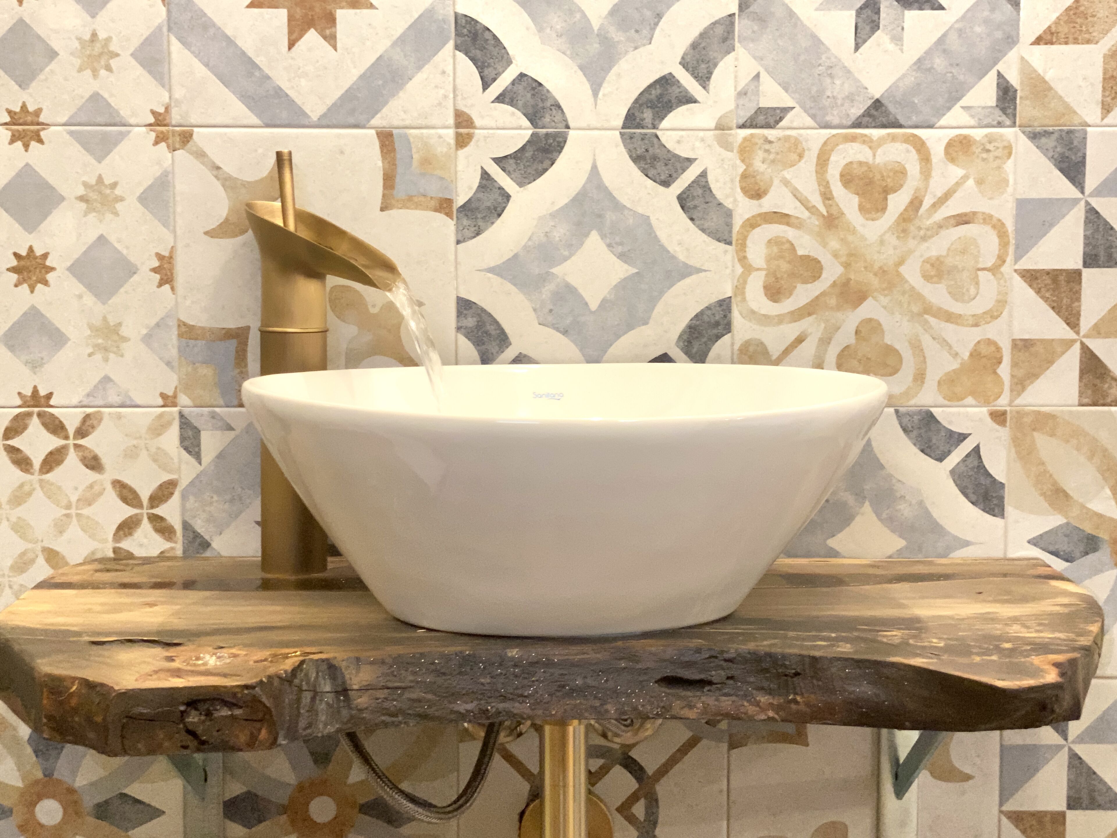 A white vessel sink mounted on a rustic wooden platform with a gold faucet, set against a tiled wall with geometric patterns.