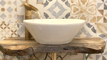 A white vessel sink mounted on a rustic wooden platform with a gold faucet, set against a tiled wall with geometric patterns.