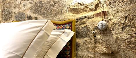 Una habitación rústica con pared de piedra, una cama con sábanas blancas, un cojín colorido, una mesita de noche de madera con una taza y un platillo, y una lámpara de pared.