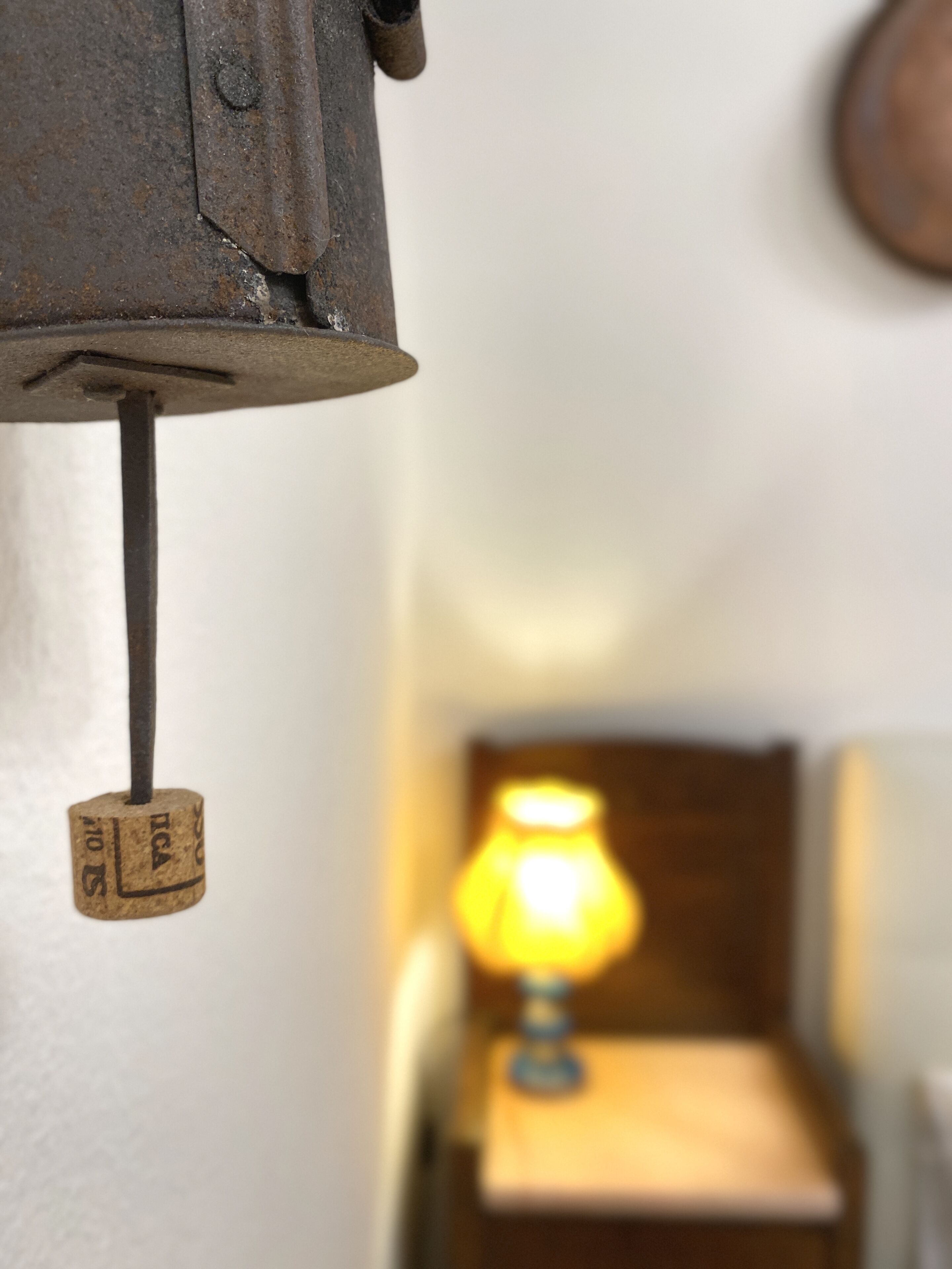 A close-up of a rustic metal bell with a cork tag hanging from it, set against a blurred background featuring a table with a lamp.