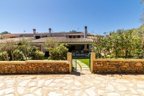 A stone-walled entrance with a gate, a paved driveway, and a building with a tiled roof and chimneys. - Villa marta, just 100 m from Cala Caterina (villasimius)