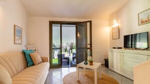 A living room with a sofa, a small table with a vase and plant, a television, and a sliding glass door leading to an outdoor area. - Villa marta, just 100 m from Cala Caterina (villasimius)