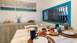 A dining area with a table set for breakfast, a view through a window with a blue frame, and artwork on the wall. - Villa marta, just 100 m from Cala Caterina (villasimius)