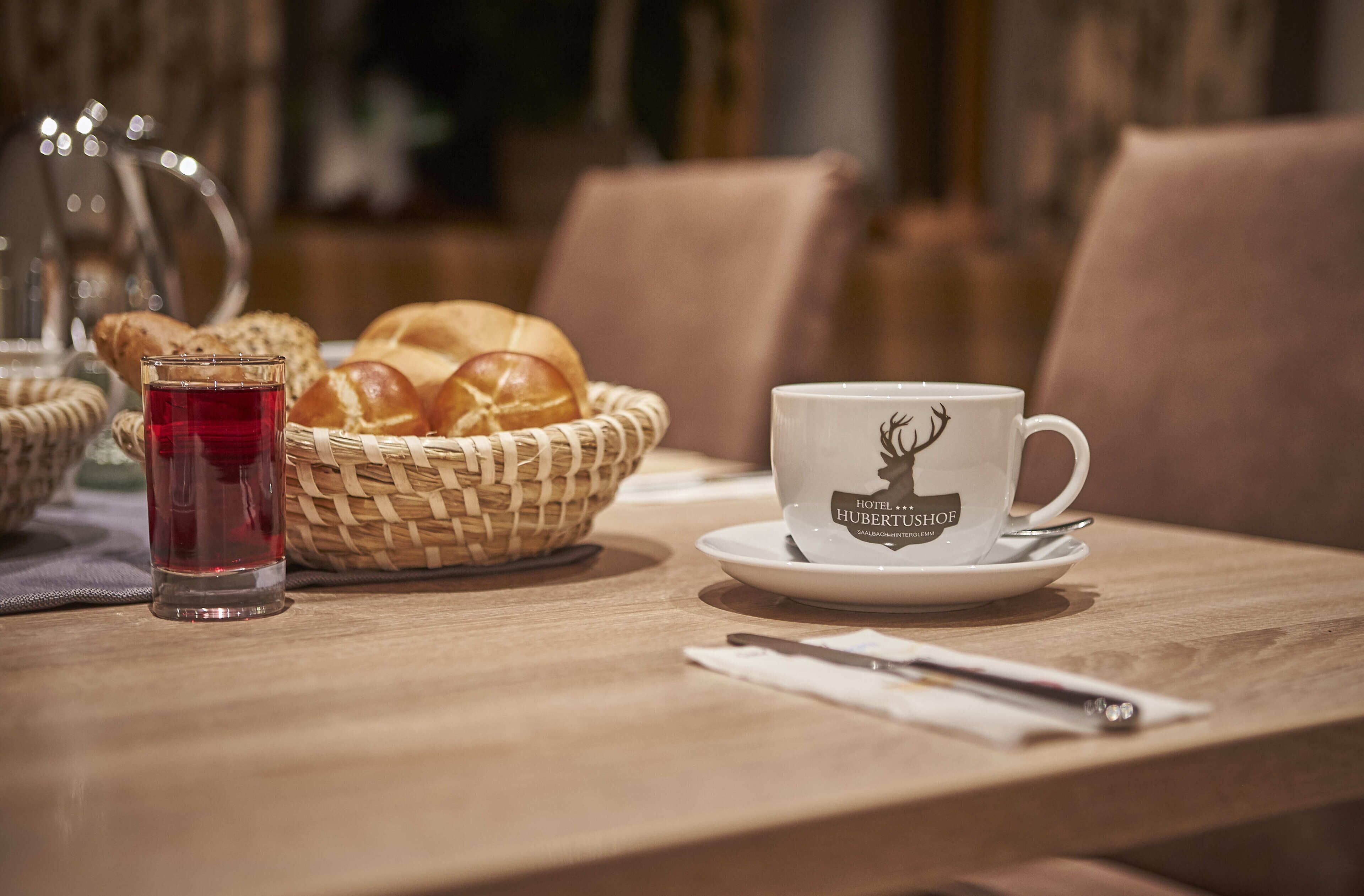 Ein Tisch mit einem Brotkorb, einem Glas roter Flüssigkeit und einer Tasse mit einem Hirsch-Logo.