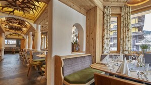 A restaurant with wooden furniture, a high ceiling with antler decorations, and a view of the outside. - Hotel Hubertushof (Saalbach-Hinterglemm)