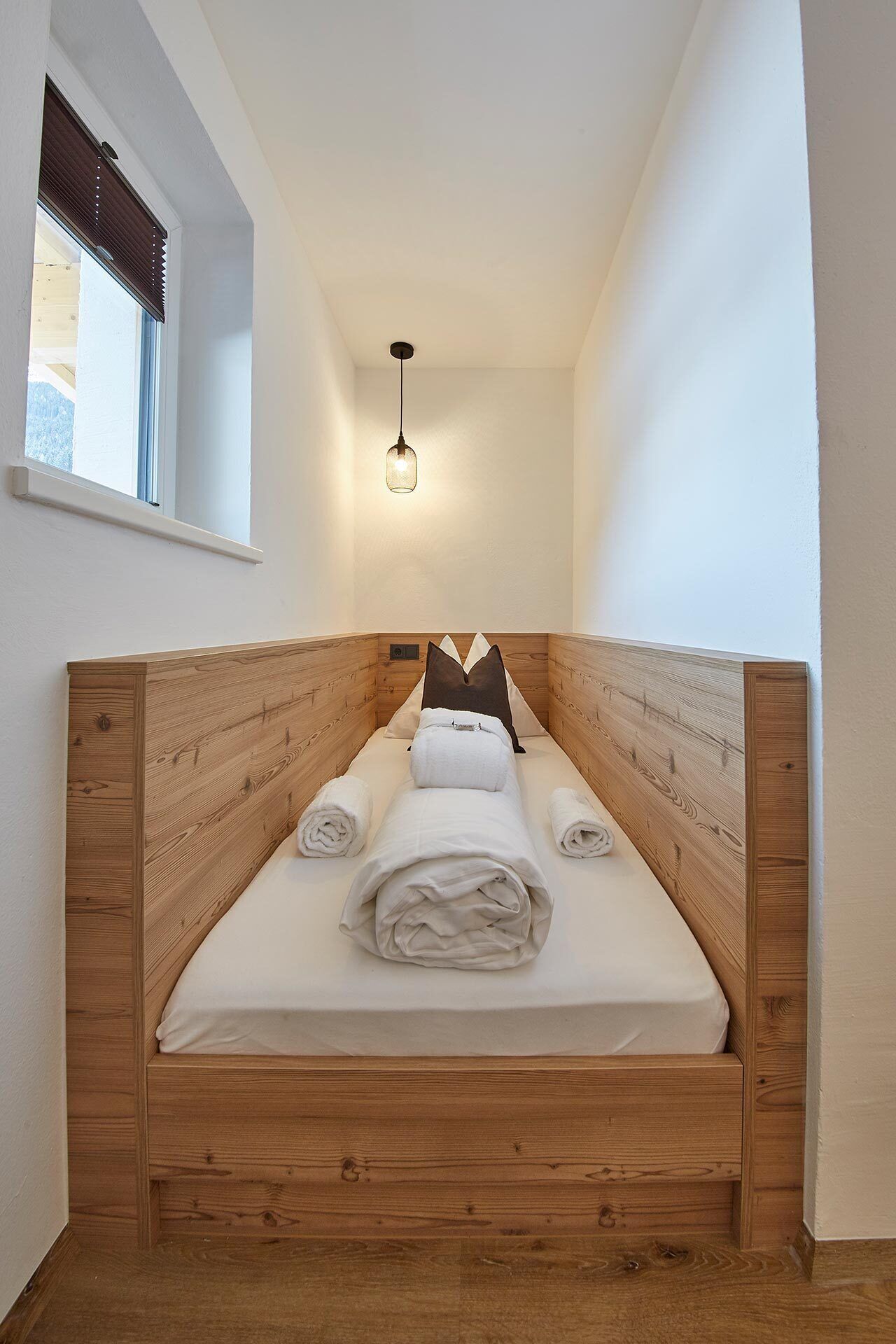 A small, single-bed room with a wooden headboard, a window with blinds, and a hanging light fixture.
