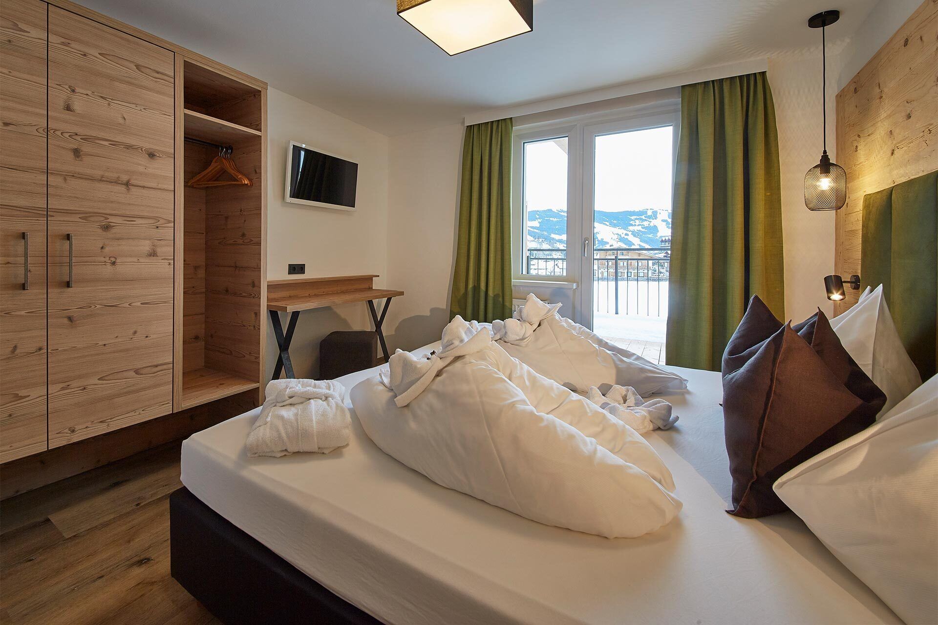 Ein Hotelzimmer mit Bett, Holzschrank, Schreibtisch, Fernseher und Bergblick durch das Fenster.