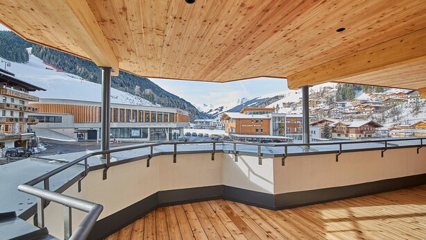 A wooden deck with a curved railing overlooking a snowy mountain town. - Hotel Hubertushof (Saalbach-Hinterglemm)