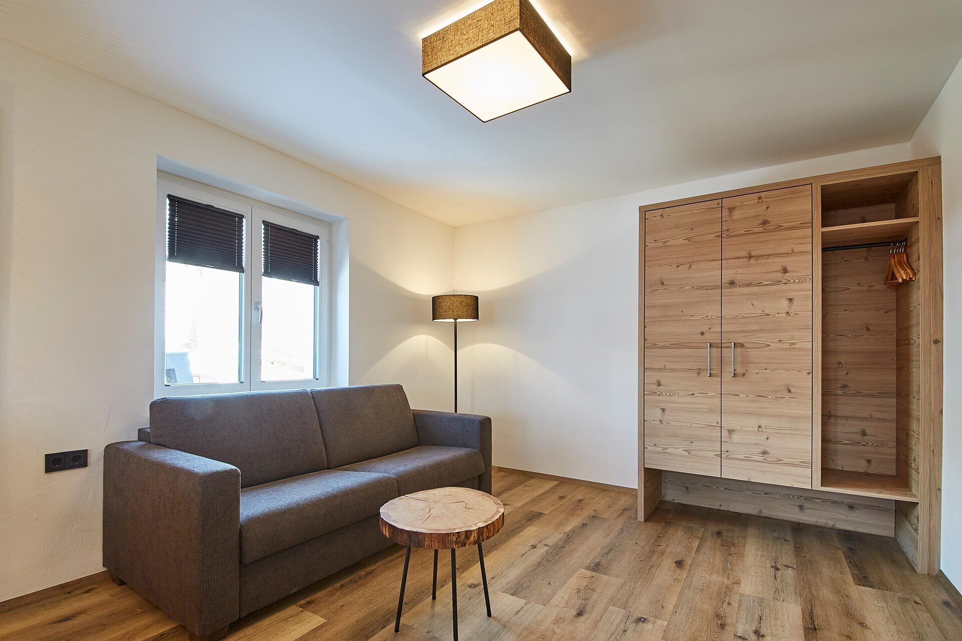 Un salon moderne avec un canapé gris, une table basse ronde en bois, un lampadaire, une armoire aux portes en bois et une fenêtre avec des stores.