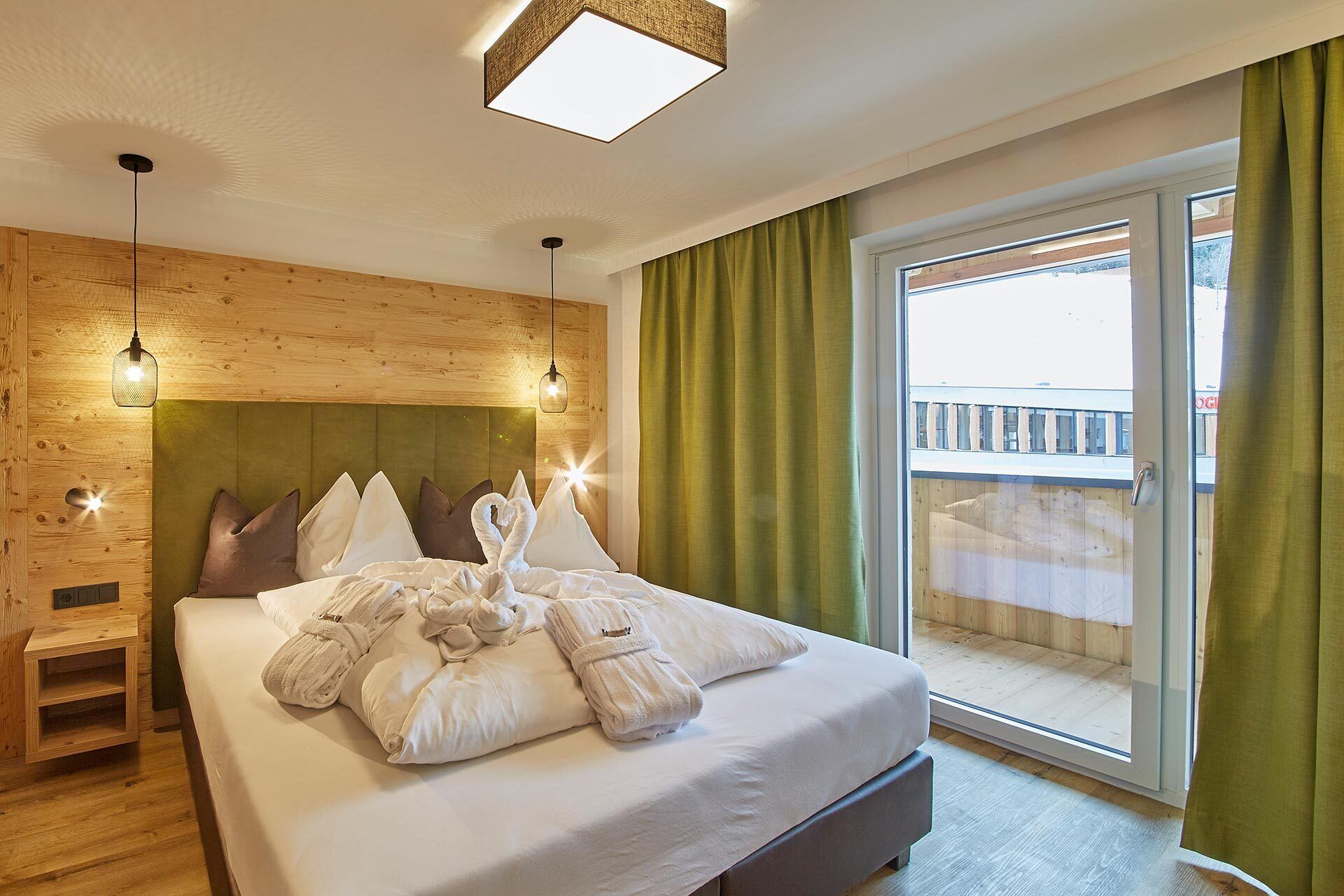Ein Hotelzimmer mit Holz-Kopfteil, einem großen Bett mit weißer Bettwäsche, einem kleinen Nachttisch und Blick auf einen Balkon durch eine Schiebetür.