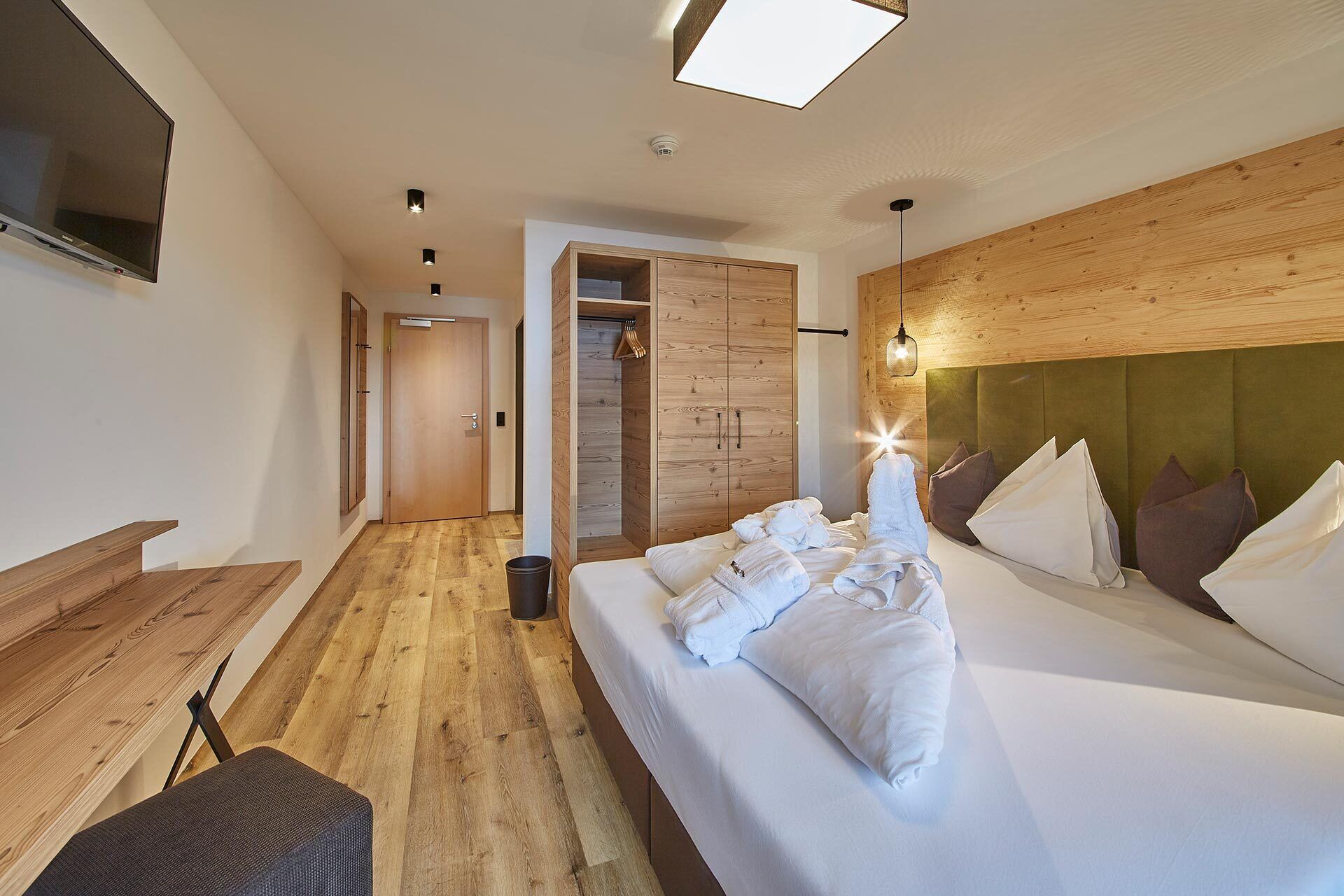 A hotel room with a wooden floor, a bed with white linens, a wooden wardrobe, and a television on the wall.