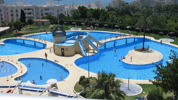 A large outdoor swimming pool with multiple sections and a central water slide, surrounded by palm trees and a view of the sea. - minerva 1 bedroom ap (Benalmádena)
