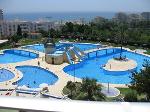 A large outdoor swimming pool with multiple sections and a central water slide, surrounded by palm trees and a view of the sea. - minerva 1 bedroom ap (Benalmádena)