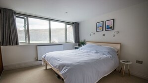 Ein Schlafzimmer mit einem großen Bett, einem Fenster mit Vorhängen, einem Heizkörper, einem Nachttisch und zwei gerahmten Bildern an der Wand.
