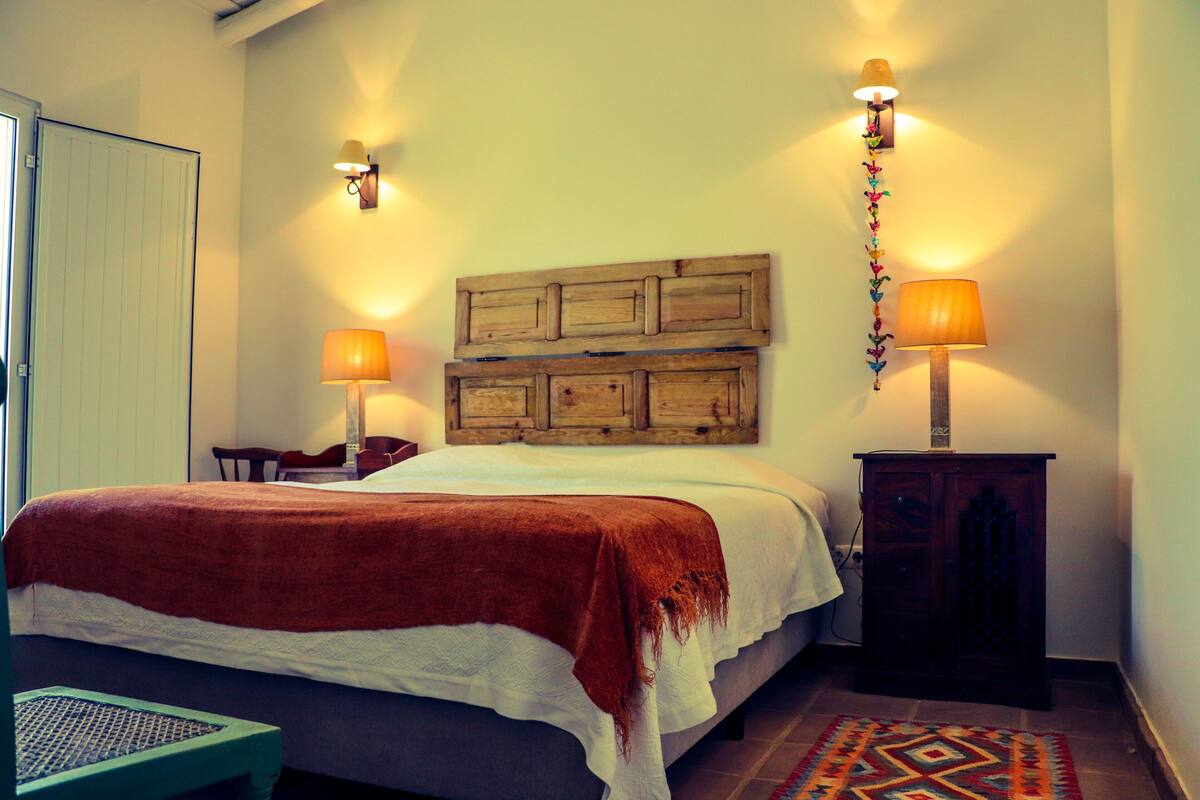 Ein Schlafzimmer mit einem Bett, zwei Wandlampen, einem Holz-Kopfteil, einer kleinen Holzkabine und einem gemusterten Teppich.