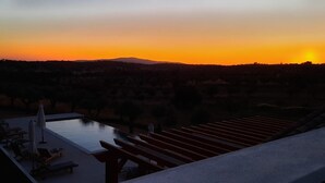 A sunset view from a balcony overlooking a pool area with lounge chairs and a pergola.