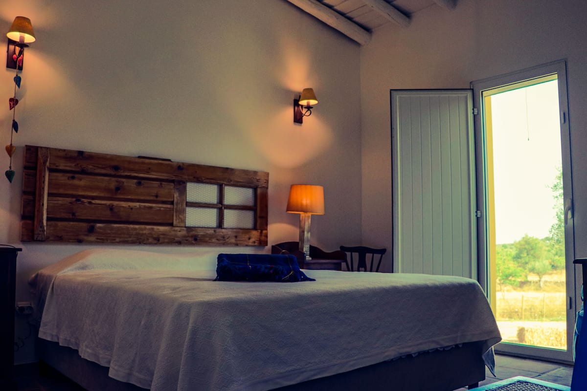 Ein Schlafzimmer mit einem hölzernen Kopfende, einem Bett mit weißer Bettdecke, einem blauen Kissen, einer Nachttischlampe und Blick ins Freie durch eine Schiebetür.
