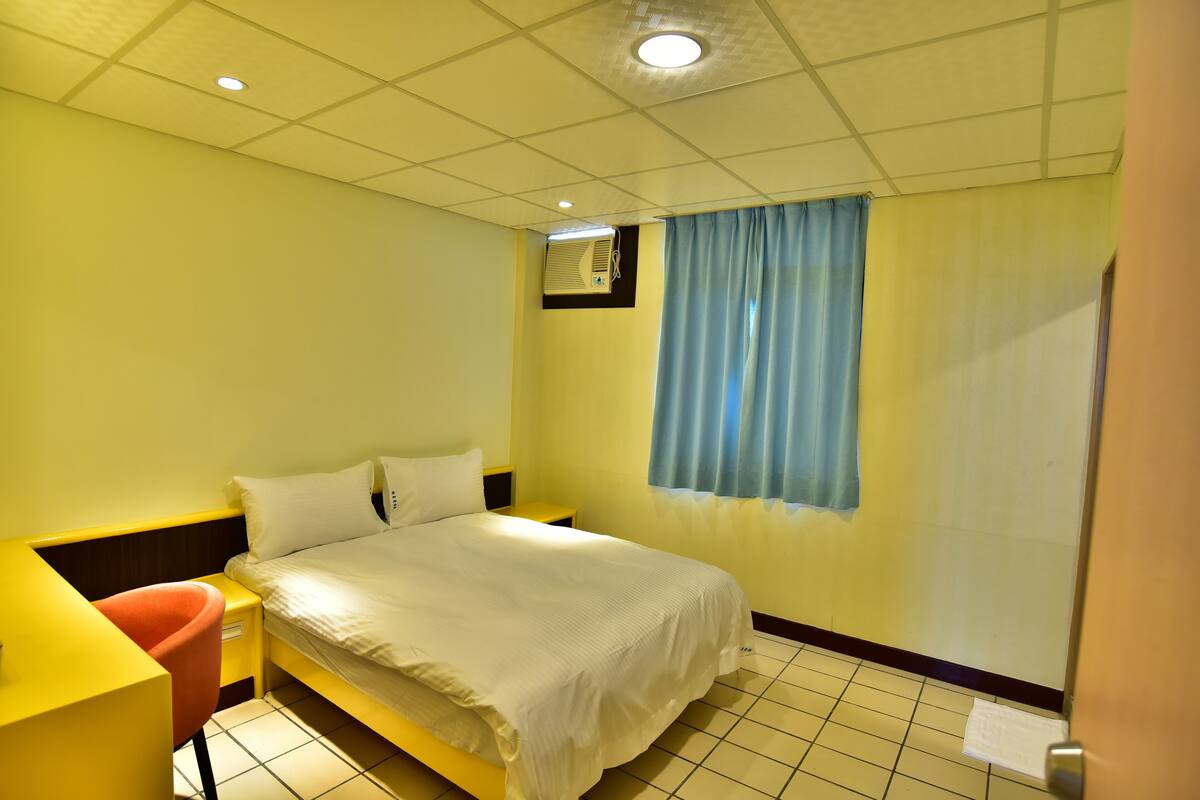 Una habitación de hotel con cama, escritorio, silla y ventana con cortinas.