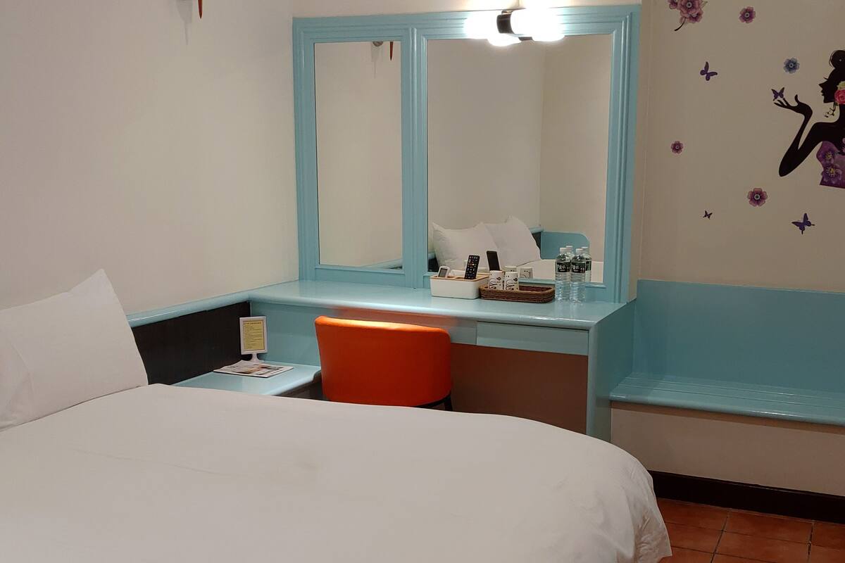 Habitación de hotel con una cama, un escritorio con silla, un espejo y una pared decorada con motivos florales.