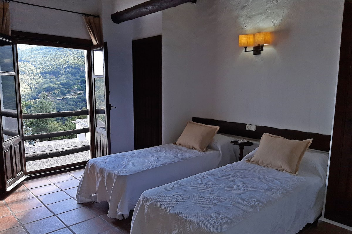 Quarto com duas camas, uma janela com vista para uma floresta e um aplique na parede.