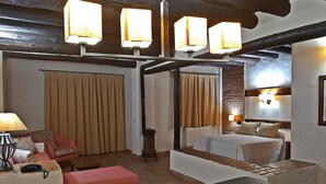 Quarto de hotel com cama, sofá, um banquinho e uma mesa com abajur. Há cortinas e uma luminária na parede.