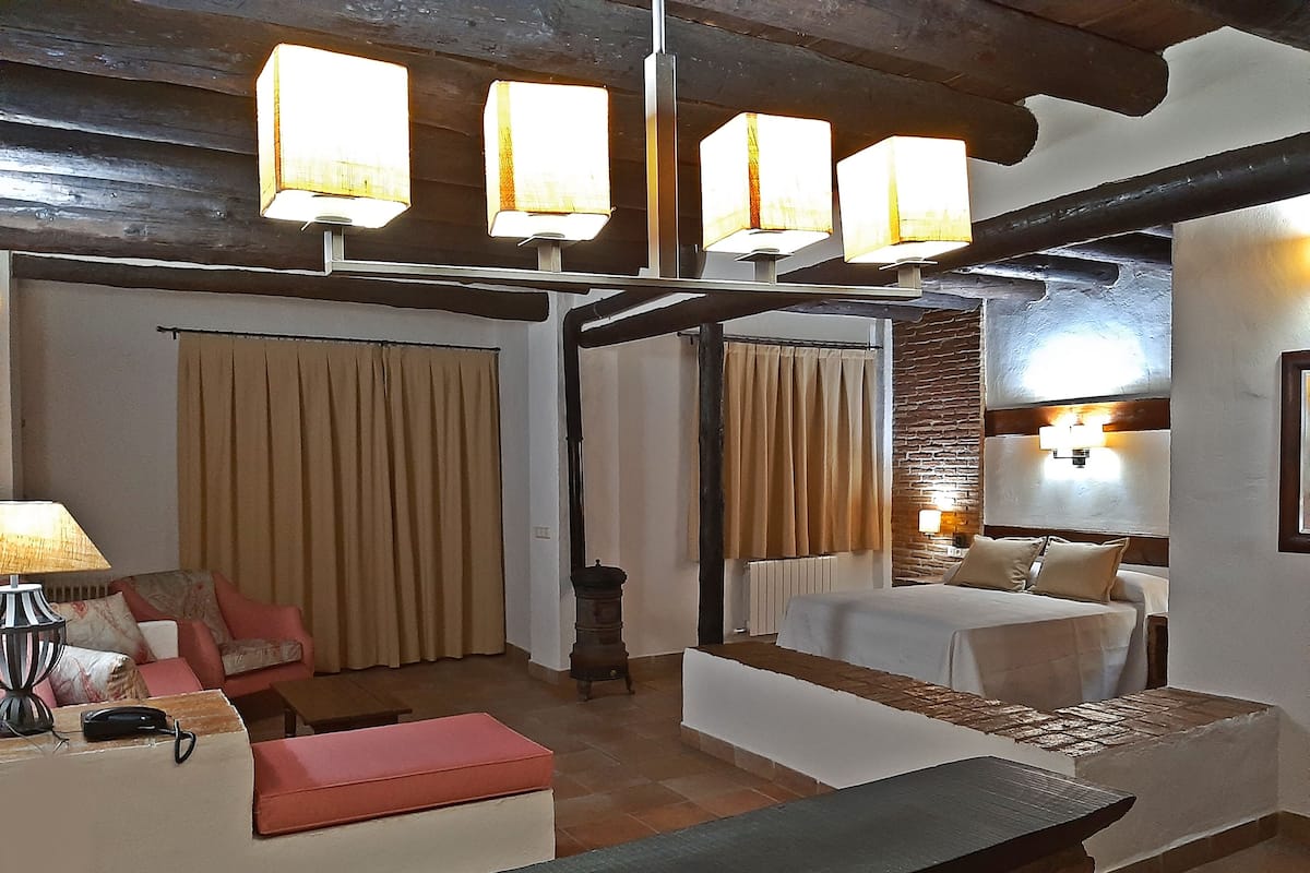 Quarto de hotel com cama, sofá, um banquinho e uma mesa com abajur. Há cortinas e uma luminária na parede.