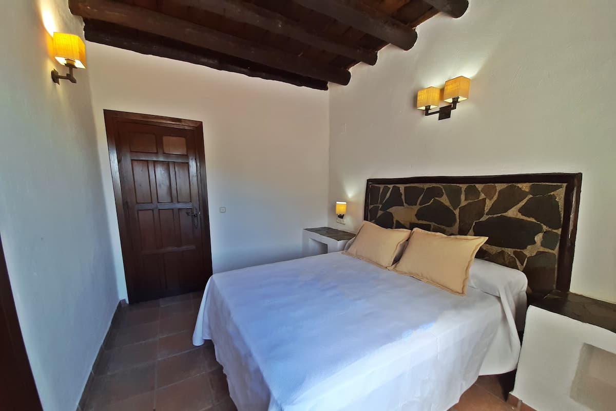 Quarto com cabeceira de pedra, porta de madeira e duas luminárias de apoio.