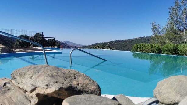 Piscina infinita azul cristalina com uma escada de metal, rodeada por pedras e árvores, com montanhas ao fundo.