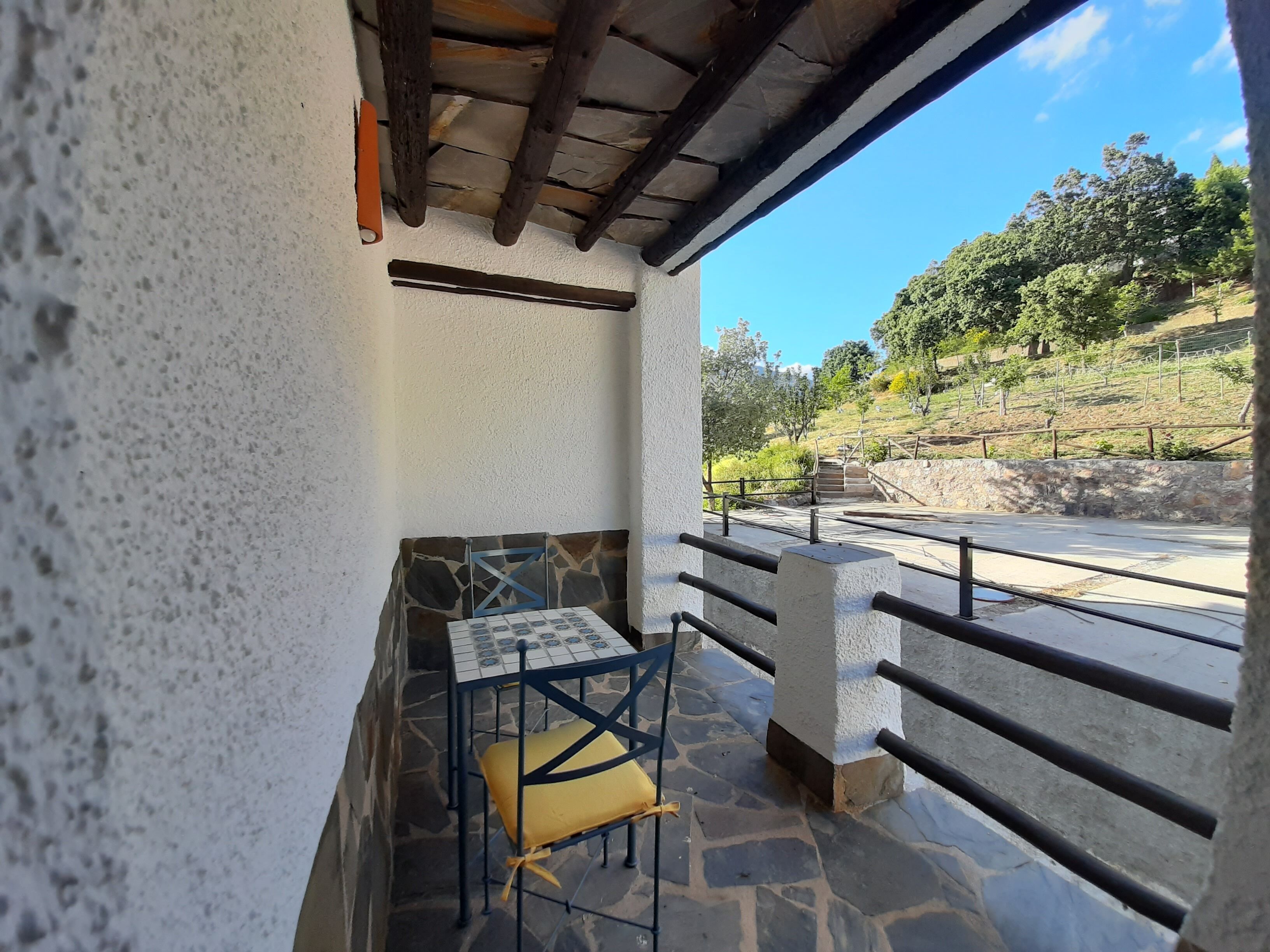 Une terrasse dallée de pierres, avec une table et des chaises, une vue sur une colline parsemée d’arbres et bordée d’une clôture, et un ciel bleu dégagé.