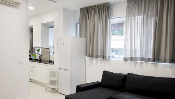 A modern living room with a black sofa, a small table, and a kitchenette in the background. - Apartamentos Funway Hostel (Madrid)