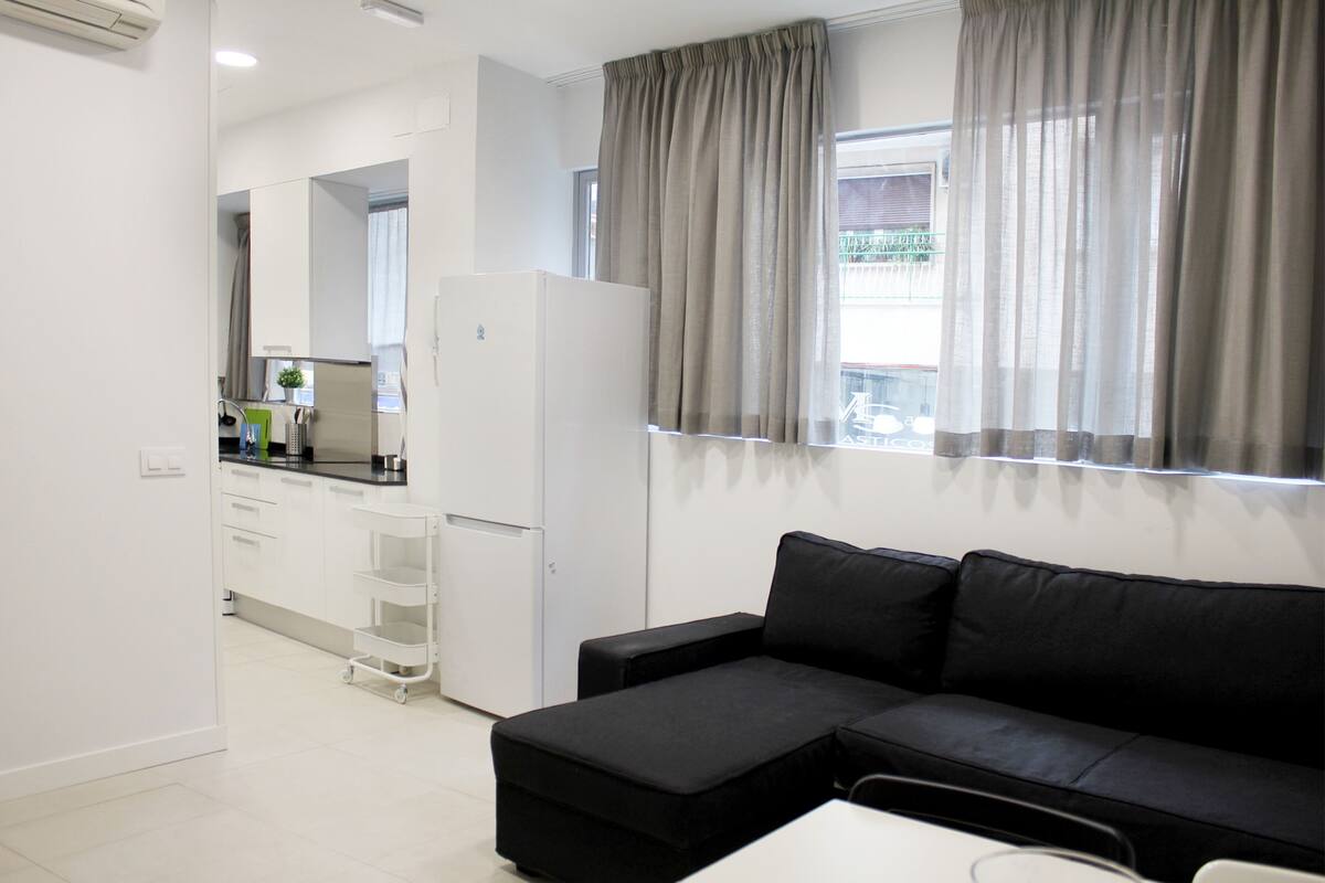 Una sala moderna con un sofá negro, una mesita y una kitchenette al fondo.