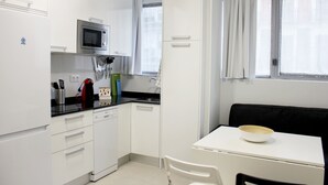 A compact kitchen with white cabinets, a microwave, and a sink. There's a dining table with a bowl on it and a black chair. - Apartamentos Funway Hostel (Madrid)