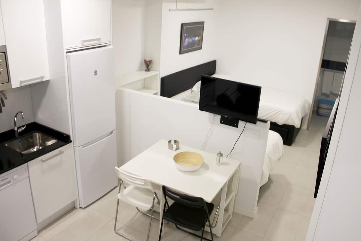 Una kitchenette compacta con refrigerador, pileta y muebles blancos. Un pequeño comedor con mesa y silla está ubicado junto a la kitchenette.