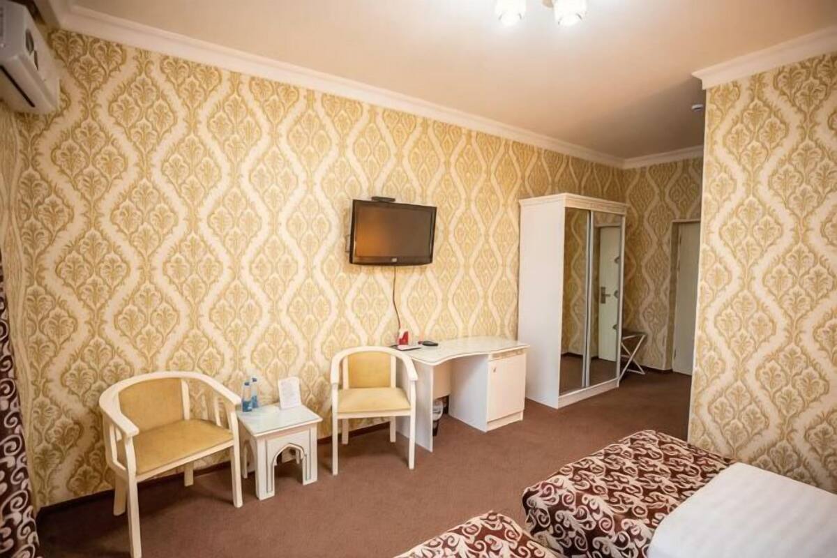 A hotel room with patterned wallpaper, a desk, two chairs, a bed, and a television.