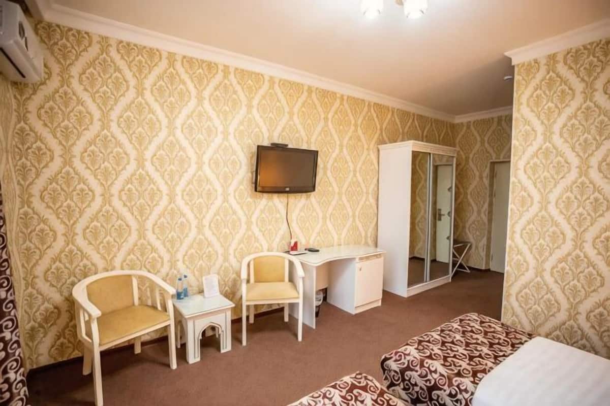 Quarto de hotel com papel de parede estampado, uma escrivaninha, duas cadeiras, uma cama e uma televisão.