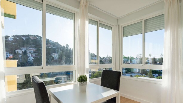 Une pièce avec une table blanche et deux chaises, de grandes fenêtres à cadres blancs, et une vue sur une colline parsemée de bâtiments.