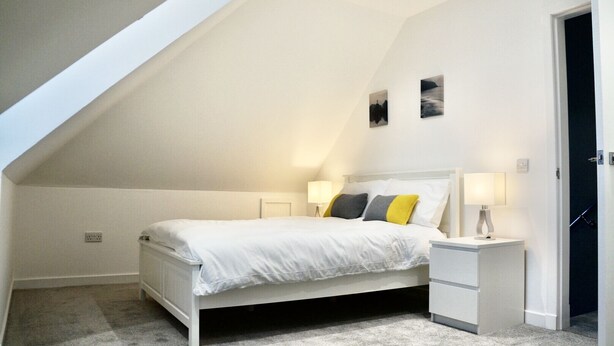 Un dormitorio con techo inclinado, una cama con ropa blanca y dos almohadas, dos lámparas de noche y una mesita de noche.