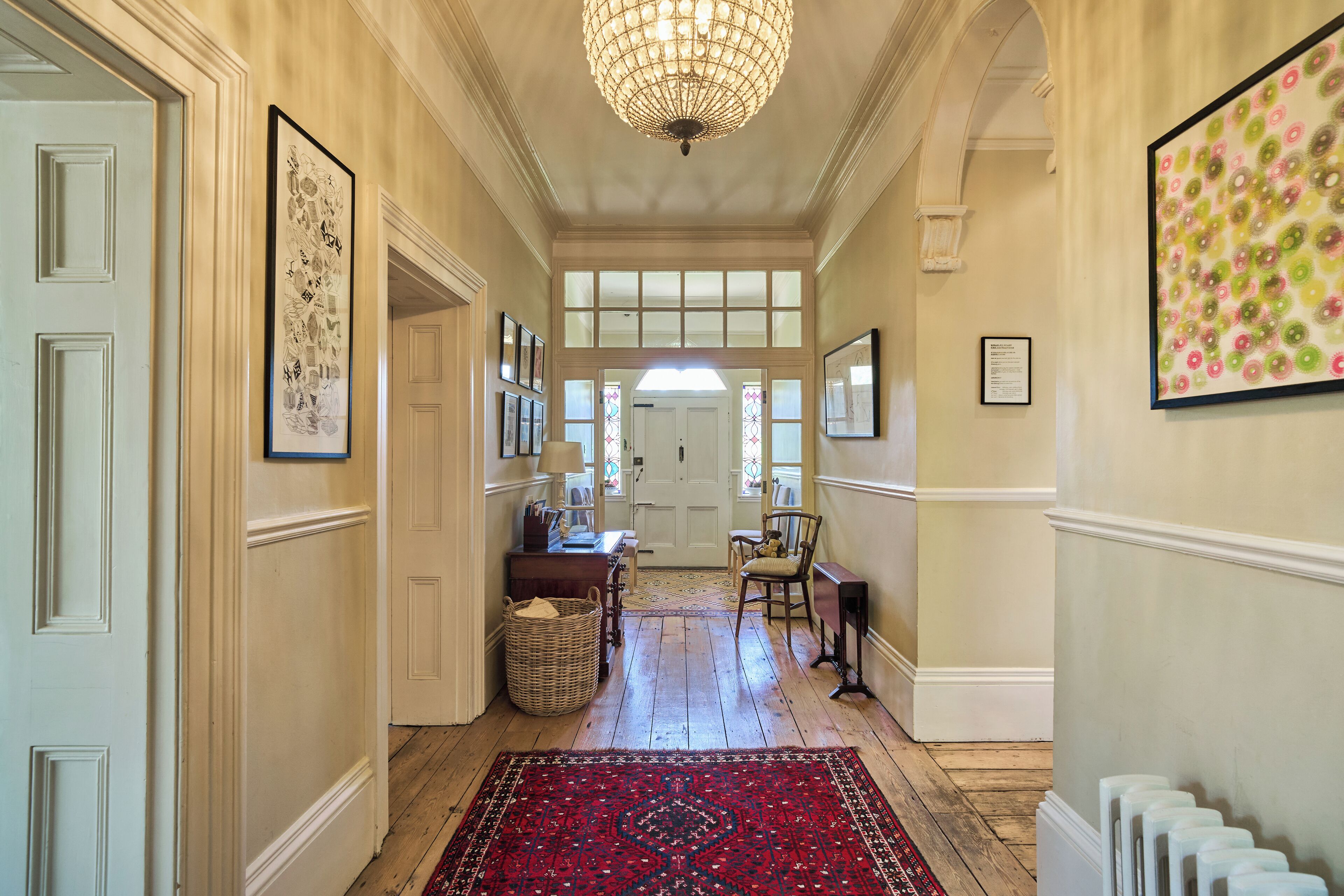 A hallway with a chandelier, framed artwork, and a red patterned rug.