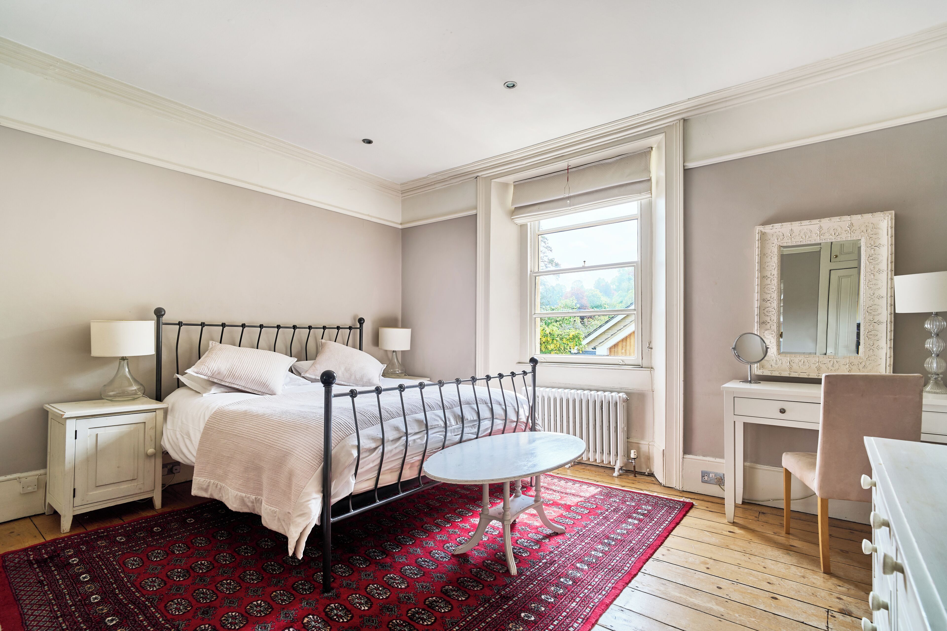Un dormitorio con cama, mesitas de noche, una pequeña mesa redonda, cómoda, espejo y ventana con vistas.