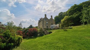 A large stone building with a prominent entrance and a covered porch, surrounded by lush green lawns and flowering shrubs. - Berkeley House (Bath)