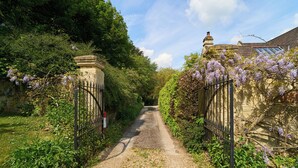 A narrow gravel path flanked by lush greenery and flowering plants, leading to a traditional house with a visible chimney and a gated entrance. - Berkeley House (Bath)