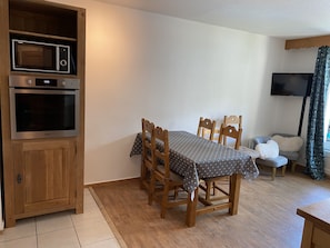 A dining area with a wooden table and chairs, a built-in microwave and oven, a television on the wall, and a patterned tablecloth.