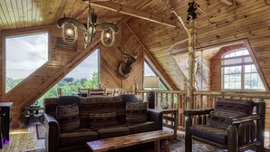 Une cabane en bois chaleureuse avec un plafond haut, une grande fenêtre, une tête de cerf accrochée au mur, ainsi qu’un canapé et un fauteuil rustiques.