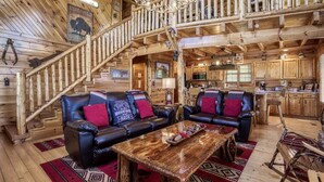 L’intérieur d’une cabane en rondins, avec un escalier en bois, des canapés en cuir, un lustre et un coin cuisine.