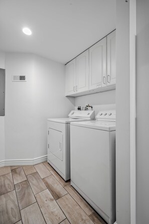 A laundry room with two front-loading washing machines and a dryer, white cabinets above, and a vent on the wall.