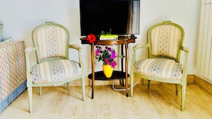 Une pièce comprenant deux chaises de style vintage, une table en bois ornée d’un vase de fleurs et un téléviseur posé sur un meuble.