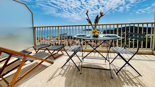 Un balcon avec une table dressée pour deux personnes, une chaise et une vue sur la mer et la ville en contrebas.