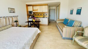 Une chambre avec un grand lit, une petite table à manger agrémentée d’un vase de fleurs, un coin cuisine et un canapé avec des coussins à motifs.